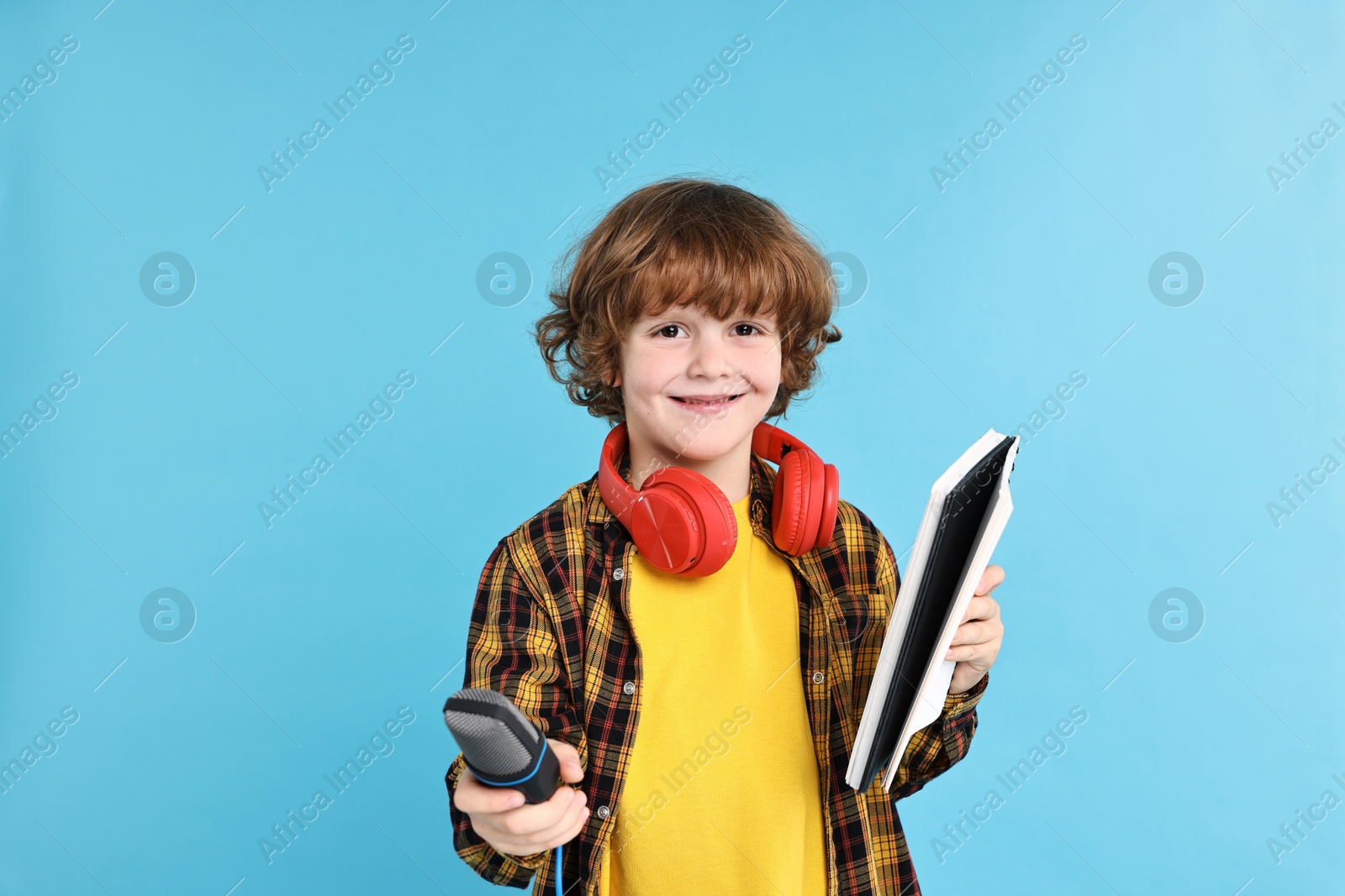 Photo of Little boy with microphone, headphones and notebook on light blue background