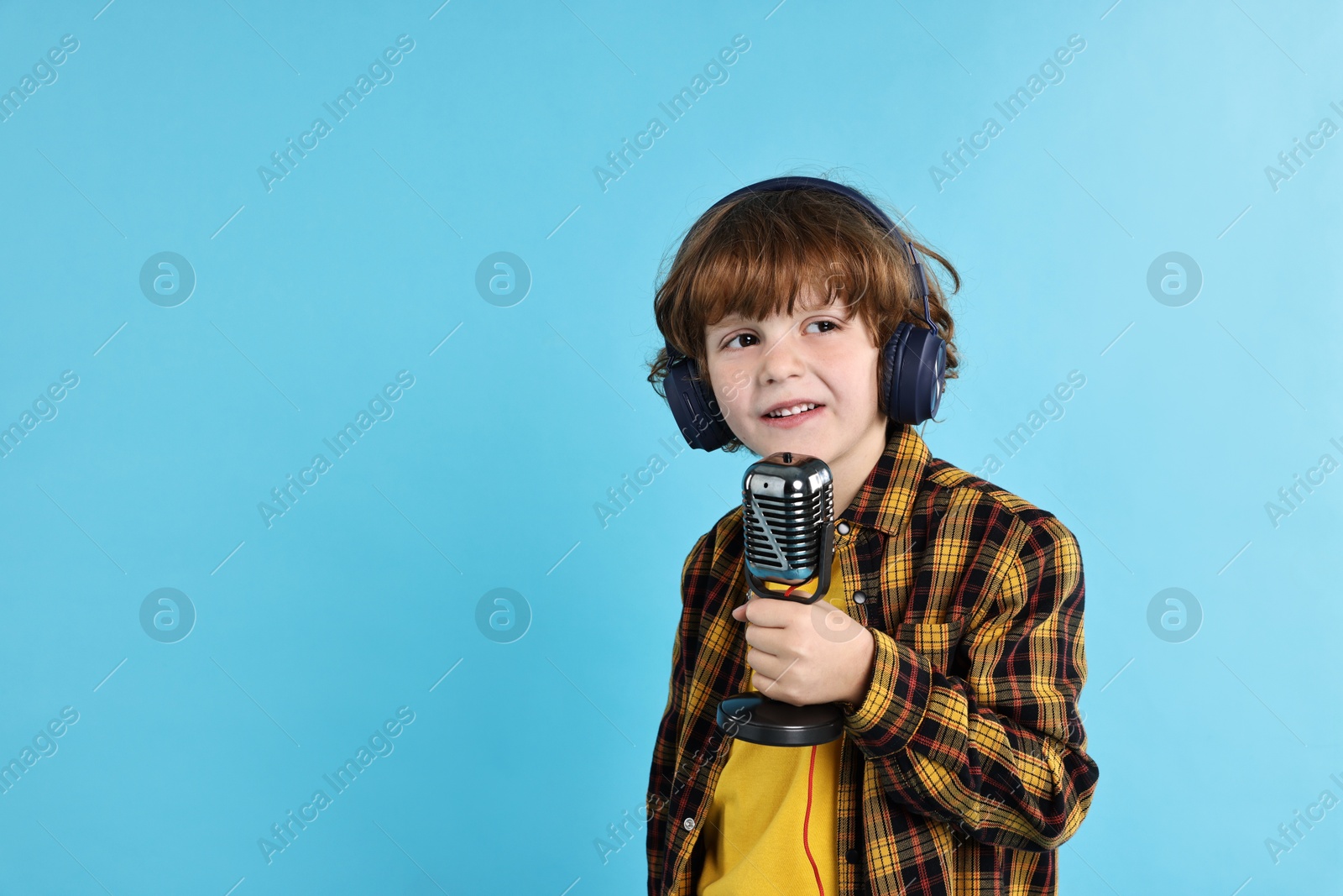 Photo of Little boy with microphone and headphones on light blue background, space for text