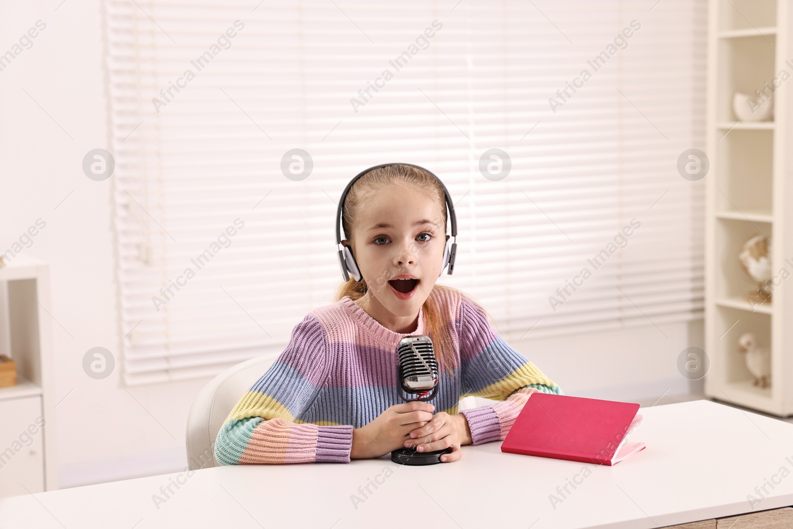 Photo of Little girl with microphone and headphones at white table indoors