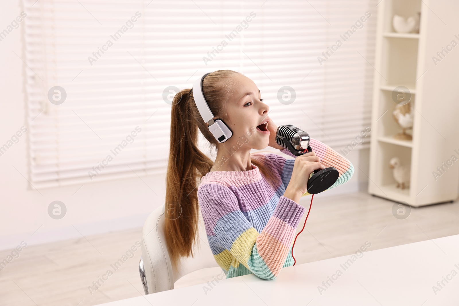 Photo of Little girl with microphone and headphones at white table indoors