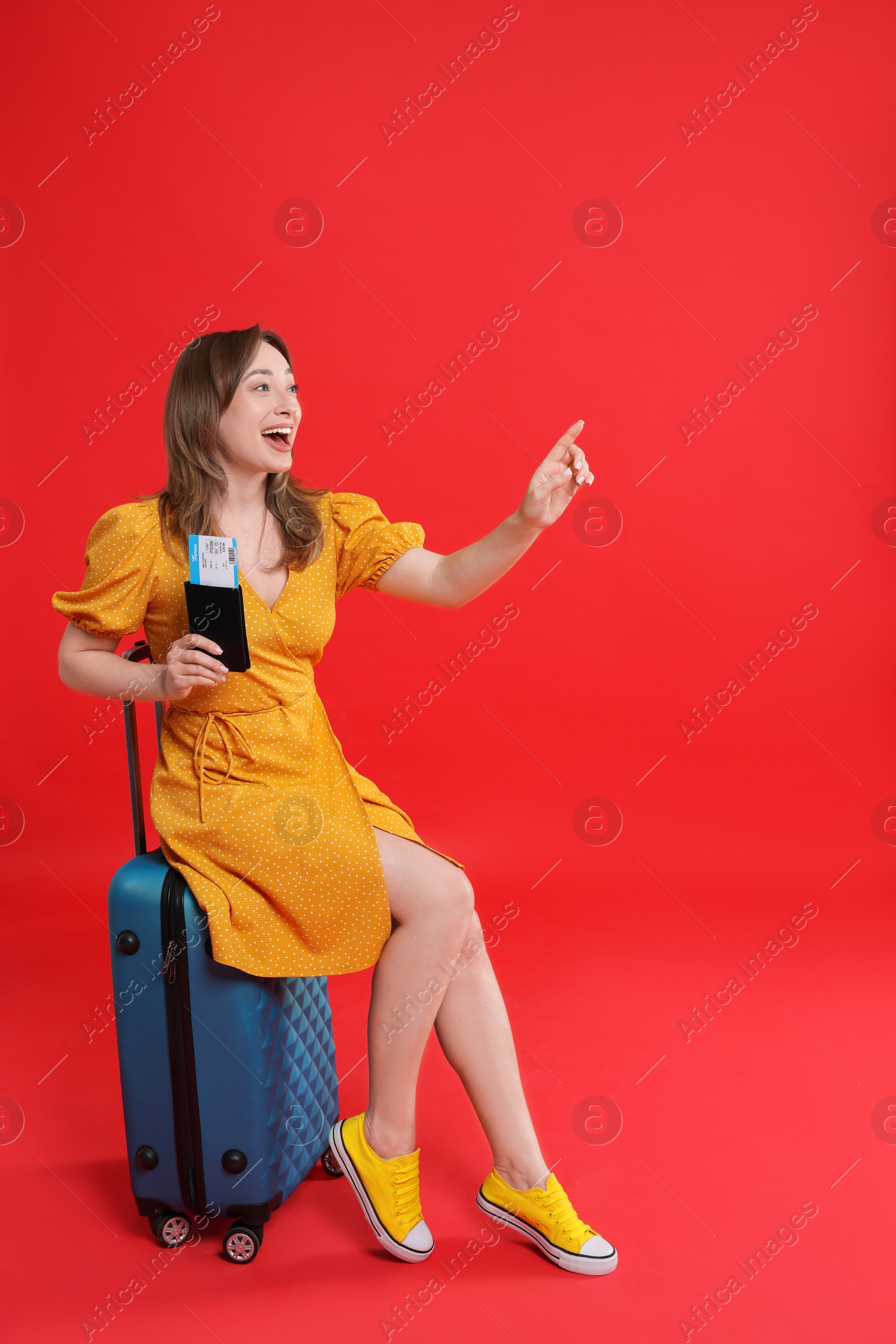 Photo of Happy traveller with passport and ticket sitting on suitcase against red background