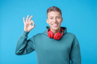 Photo of Happy young man with headphones showing OK gesture on light blue background