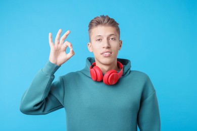 Young man with headphones showing OK gesture on light blue background