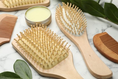 Photo of Wooden hair brushes, combs, wax and green leaves on white marble table, closeup