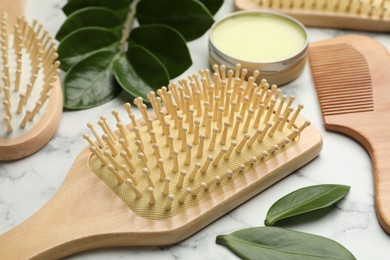 Photo of Wooden hair brushes, comb, wax and green leaves on white marble table, closeup