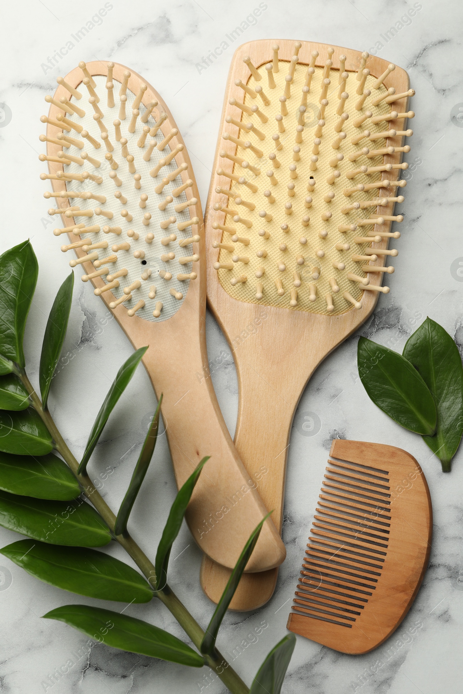 Photo of Wooden hair brushes, comb and green leaves on white marble table, flat lay