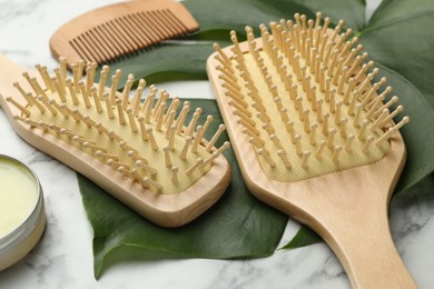 Photo of Wooden hair brushes, comb, wax and green leaves on white marble table, closeup