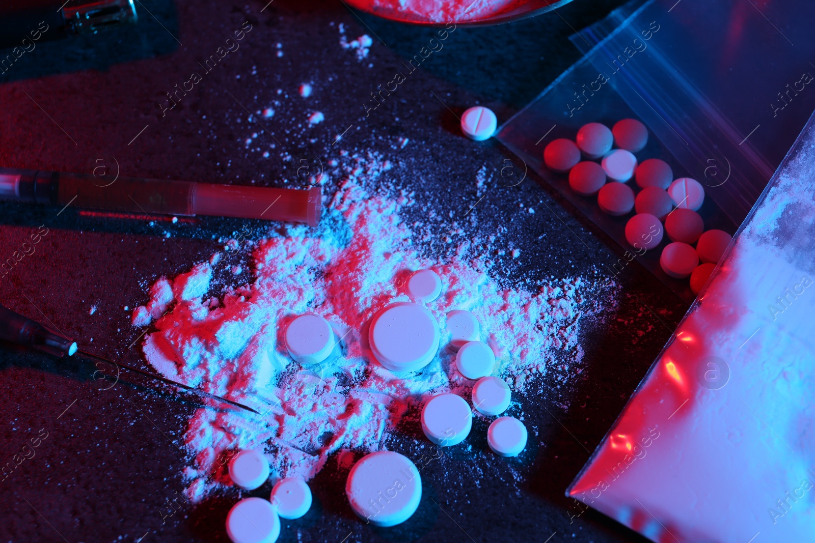 Photo of Drug addiction. Plastic bag with powder, pills and syringes on dark table in neon lights, flat lay