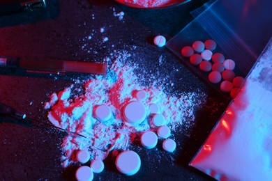 Photo of Drug addiction. Plastic bag with powder, pills and syringes on dark table in neon lights, flat lay