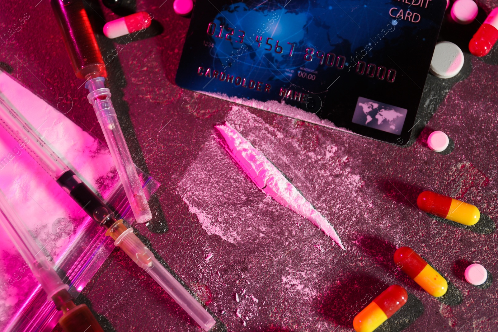 Photo of Drug addiction. Plastic bag with powder, credit card, pills and syringes on dark textured table in color light, flat lay