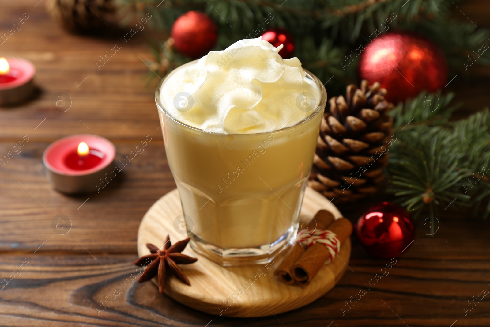 Photo of Tasty Christmas cocktail (eggnog) in glass, festive decor and burning candles on wooden table, closeup