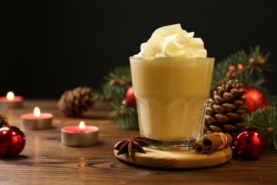 Photo of Tasty Christmas cocktail (eggnog) in glass, festive decor and burning candles on wooden table, closeup