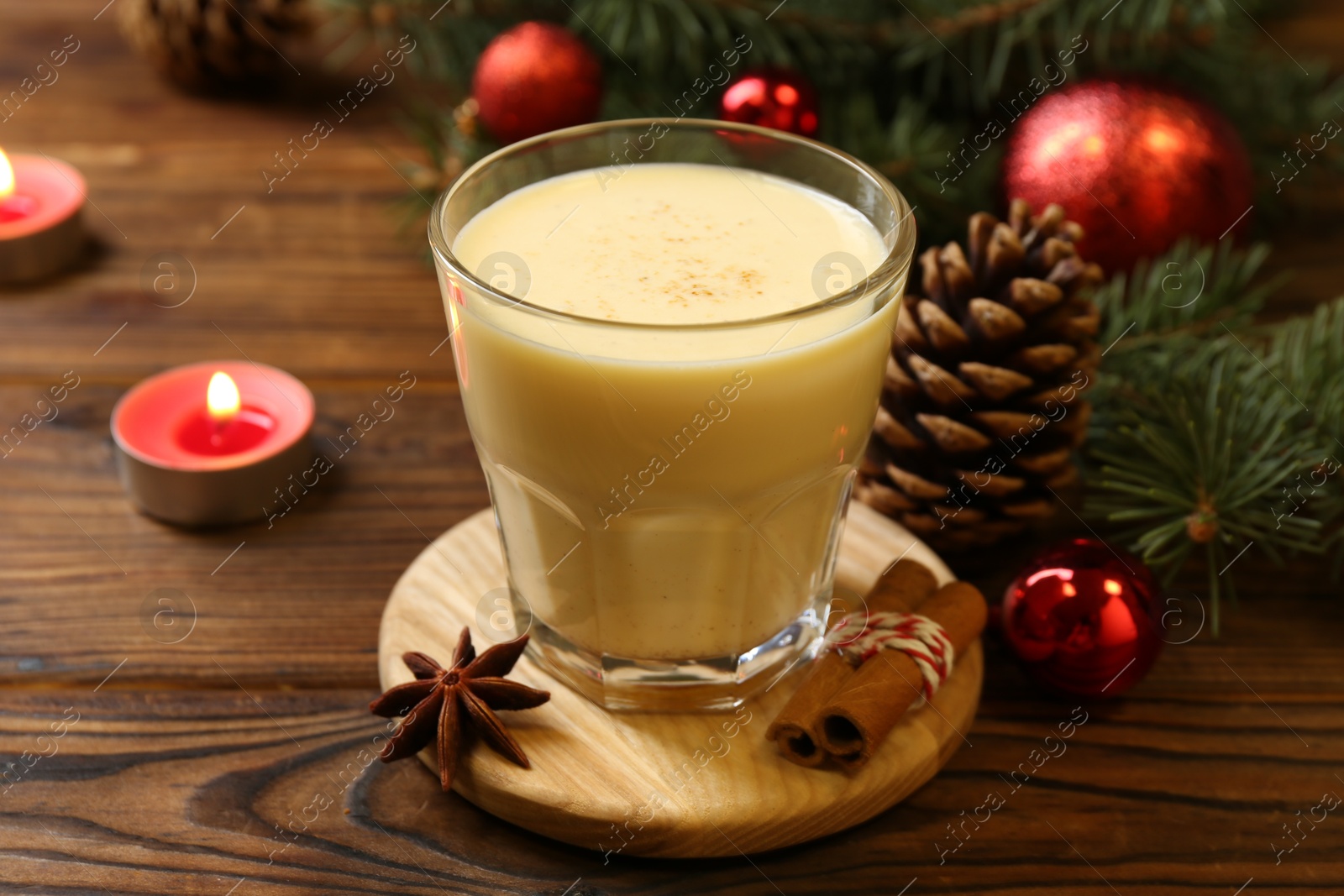 Photo of Tasty Christmas cocktail (eggnog) in glass, festive decor and burning candles on wooden table, closeup