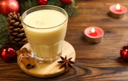 Photo of Tasty Christmas cocktail (eggnog) in glass, festive decor and burning candles on wooden table, closeup