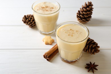 Photo of Tasty Christmas cocktail (eggnog) in glasses, pine cones and spices on white wooden table, closeup. Space for text