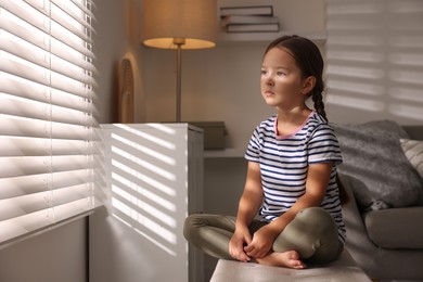 Photo of Orphanage concept. Sad girl sitting on bench and looking through window blinds indoors