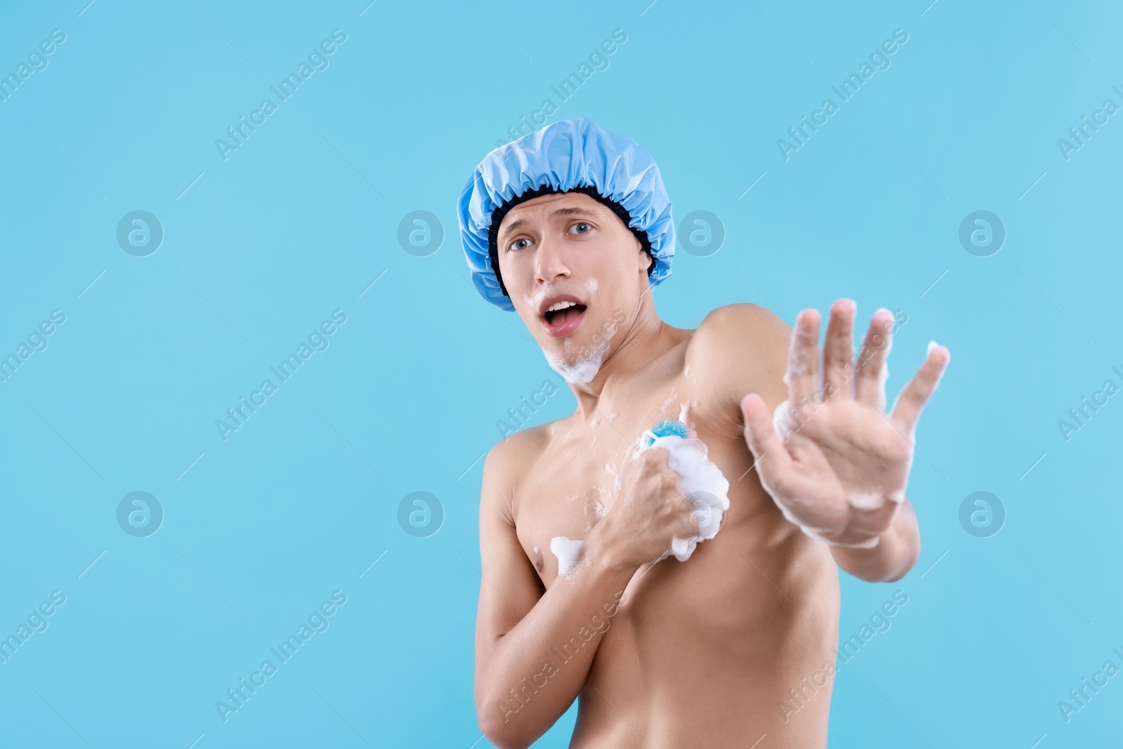 Photo of Emotional man with shower cap and mesh sponge making stop gesture on light blue background, space for text