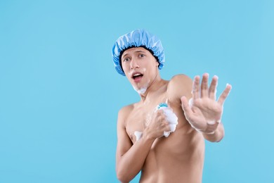 Photo of Emotional man with shower cap and mesh sponge making stop gesture on light blue background, space for text