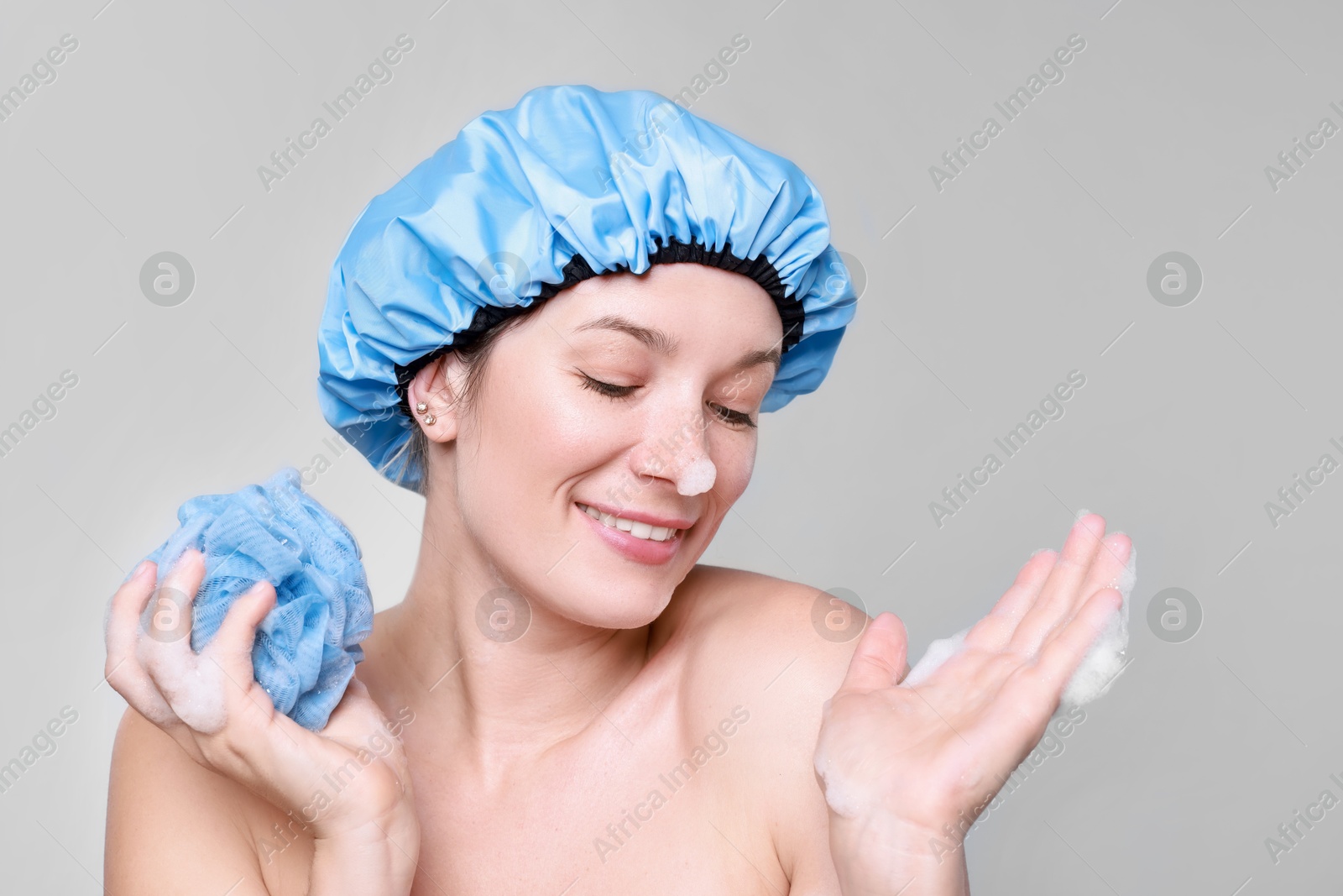 Photo of Woman in shower cap covered with soap foam holding mesh sponge on grey background