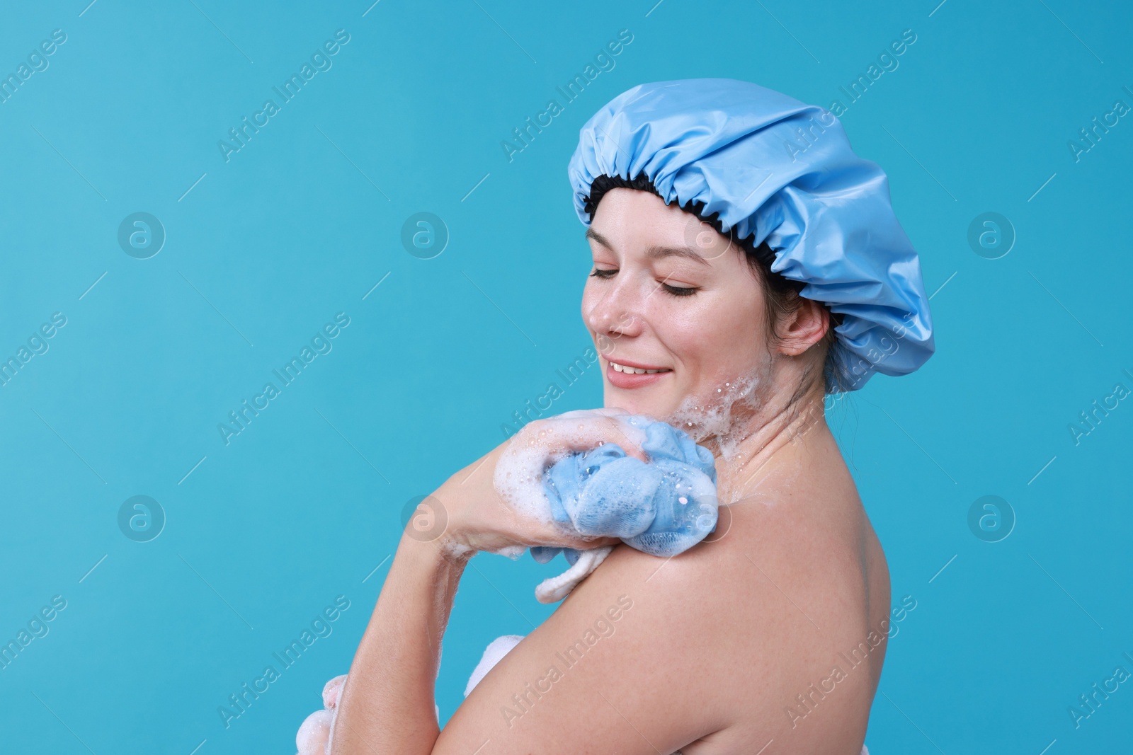 Photo of Woman in shower cap covered with soap foam holding mesh sponge on light blue background