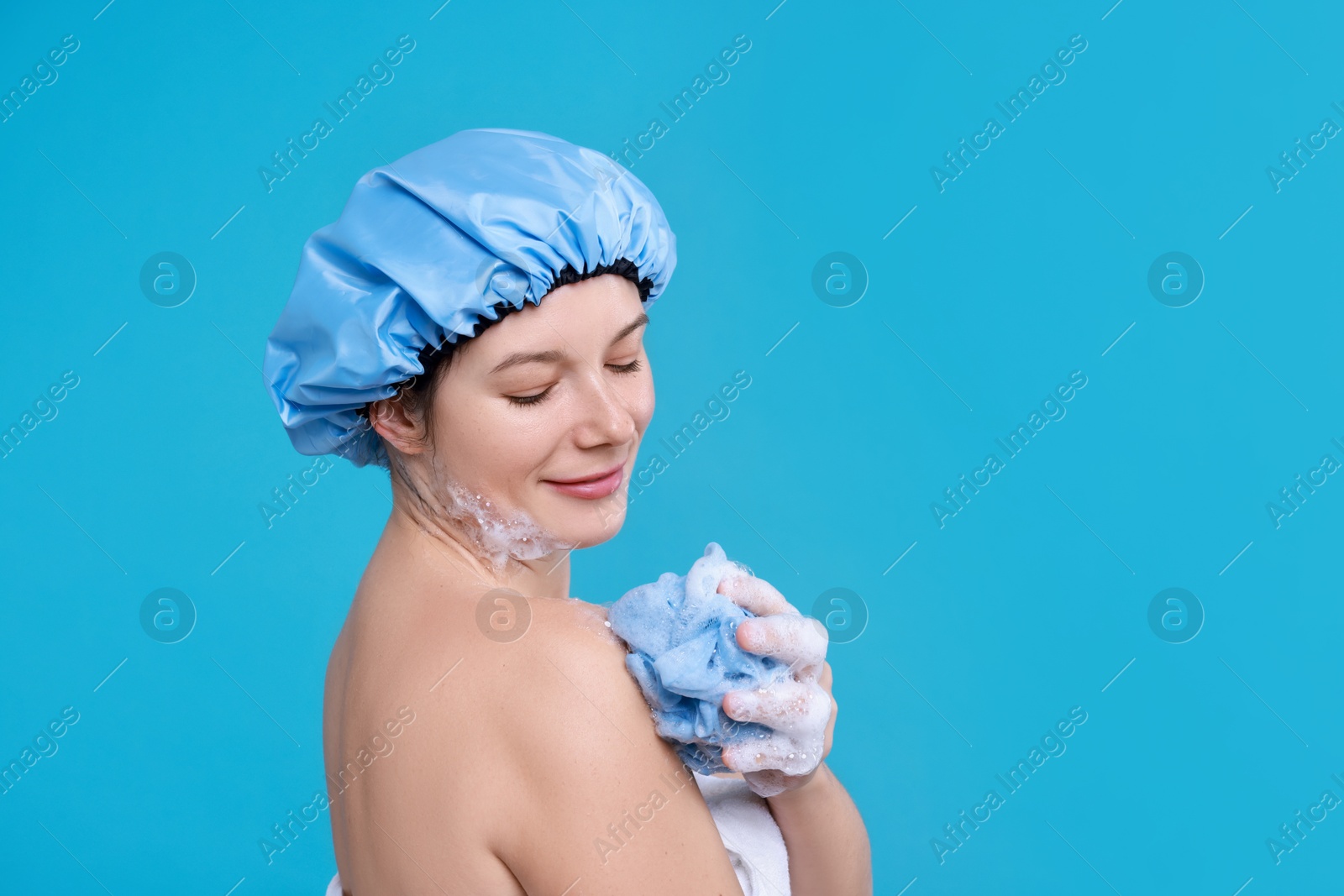 Photo of Woman in shower cap covered with soap foam holding mesh sponge on light blue background