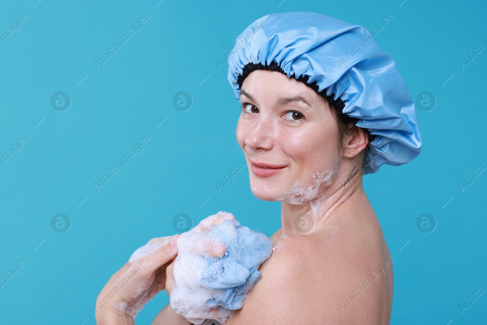 Photo of Woman in shower cap covered with soap foam holding mesh sponge on light blue background