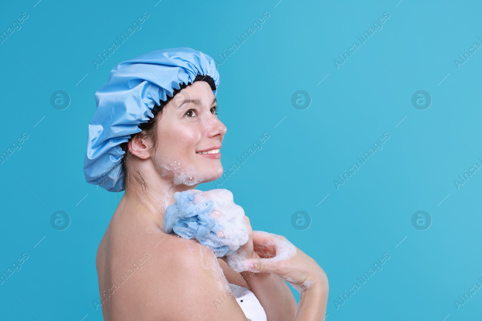 Photo of Woman in shower cap covered with soap foam holding mesh sponge on light blue background, space for text