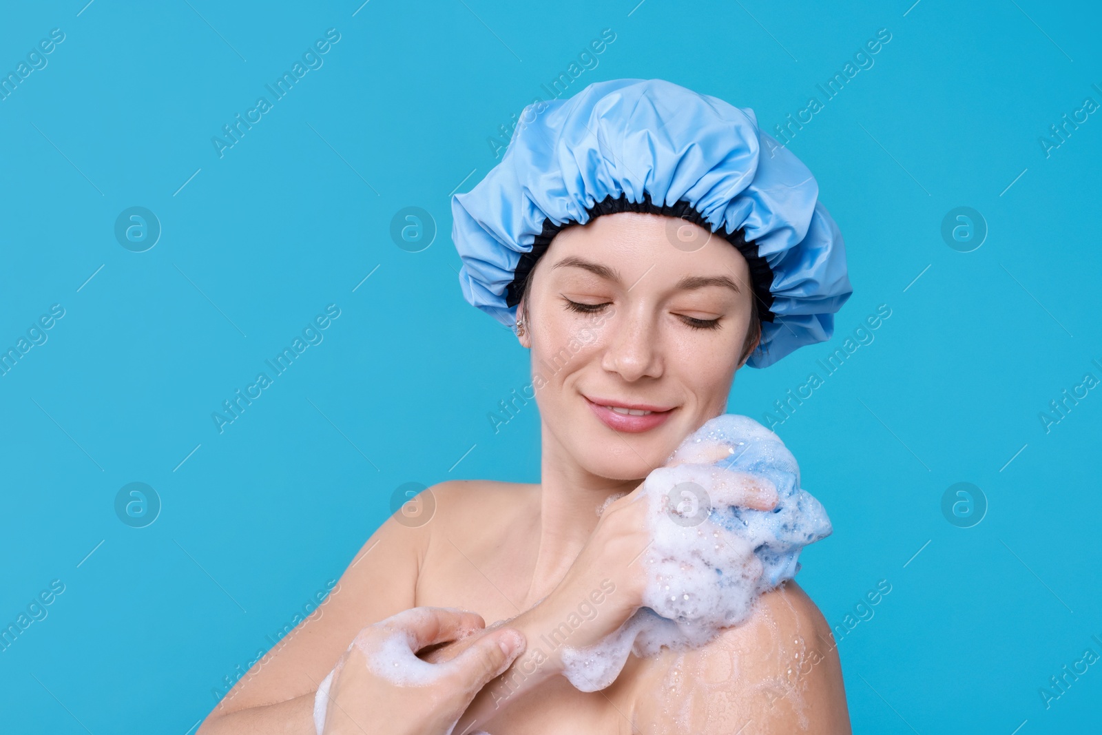 Photo of Woman in shower cap covered with soap foam holding mesh sponge on light blue background