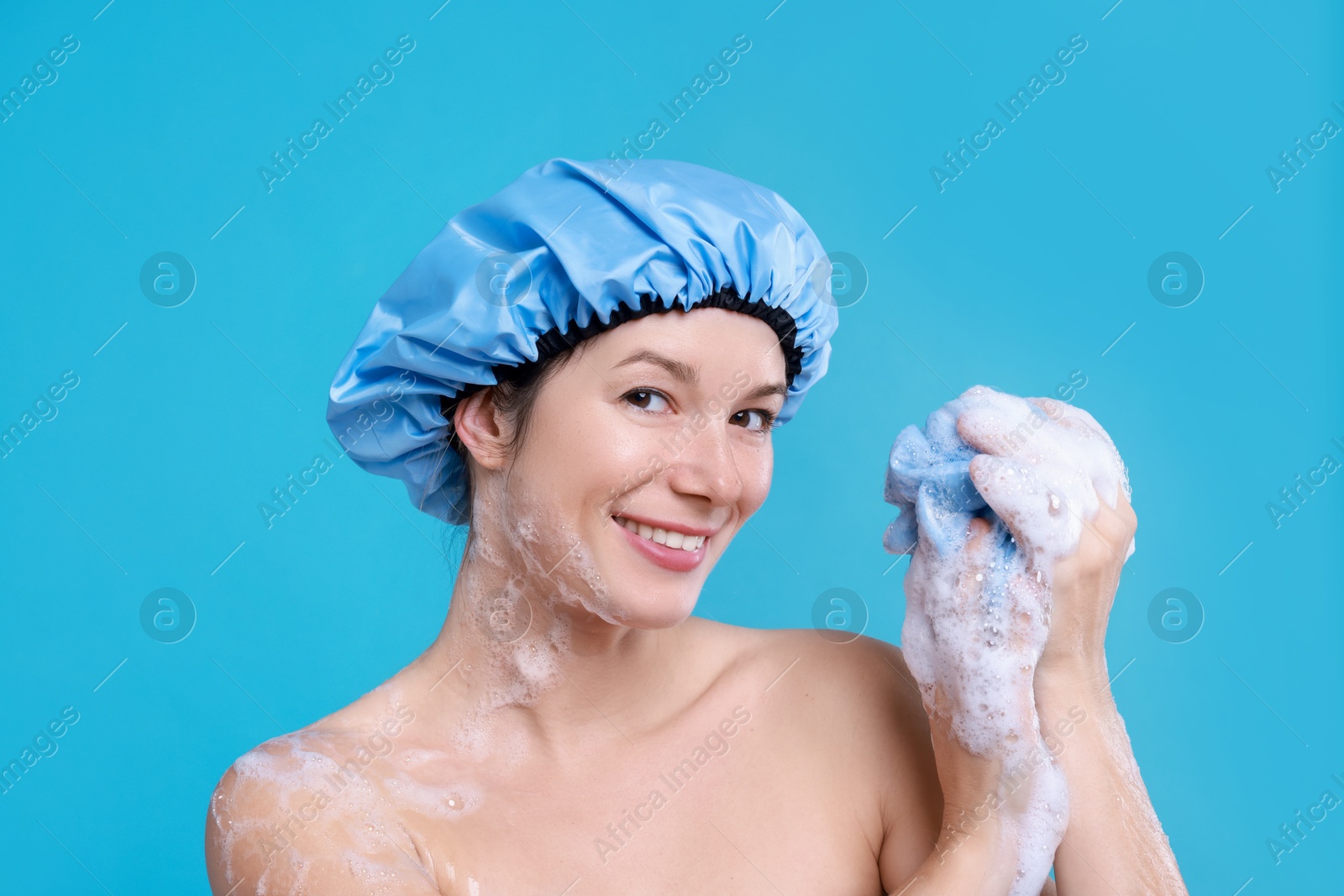 Photo of Woman in shower cap covered with soap foam holding mesh sponge on light blue background