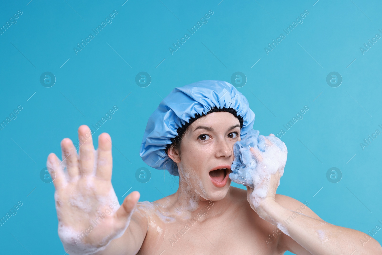 Photo of Emotional woman with shower cap and mesh sponge making stop gesture on light blue background