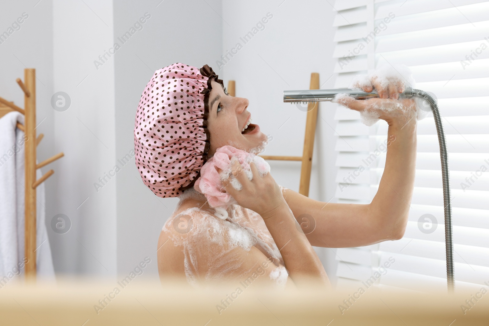 Photo of Woman with cap and mesh sponge singing while taking shower in bathroom