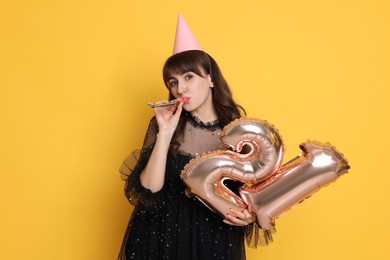 Photo of Coming of age party - 21st birthday. Young woman with hat and blower holding number shaped balloons on yellow background