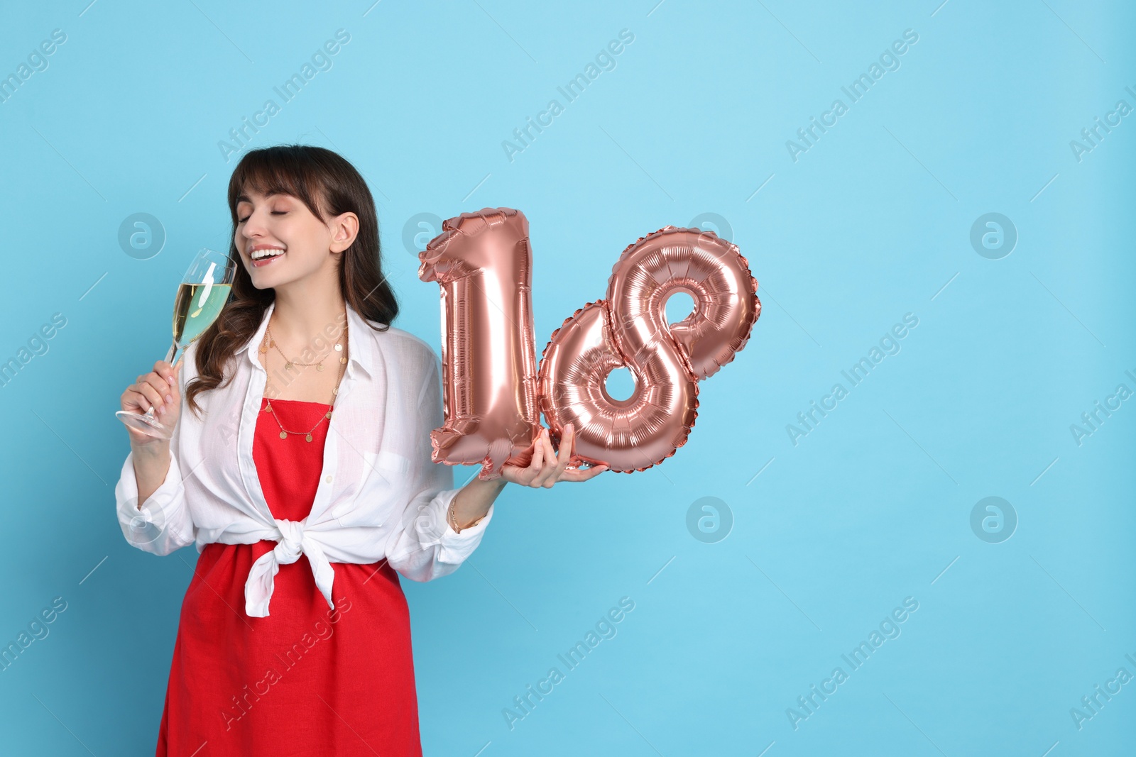 Photo of Coming of age party - 18th birthday. Young woman with glass of wine holding number shaped balloons on light blue background, space for text