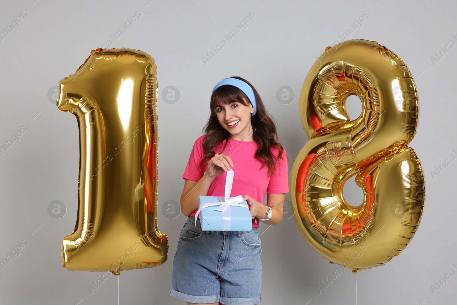 Photo of Coming of age party - 18th birthday. Young woman with gift box holding number shaped balloons on light grey background