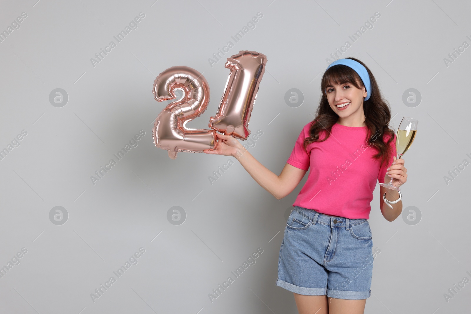 Photo of Coming of age party - 21st birthday. Young woman with glass of wine holding number shaped balloons on light grey background, space for text