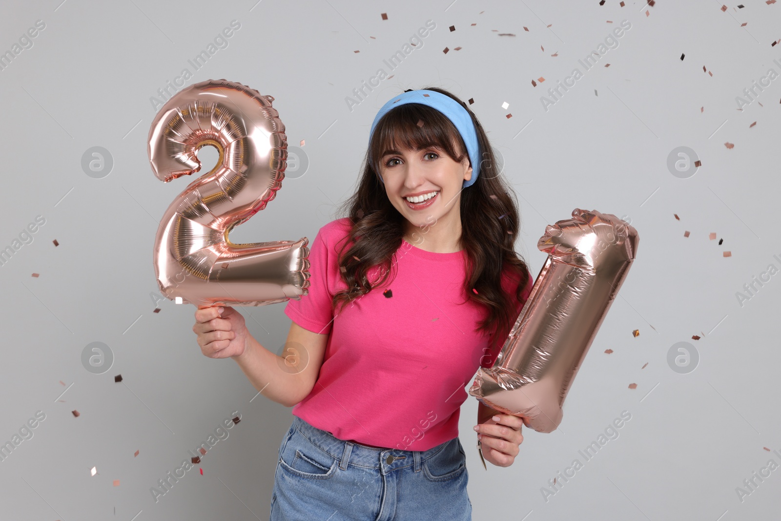 Photo of Coming of age party - 21st birthday. Young woman holding number shaped balloons on light grey background with confetti