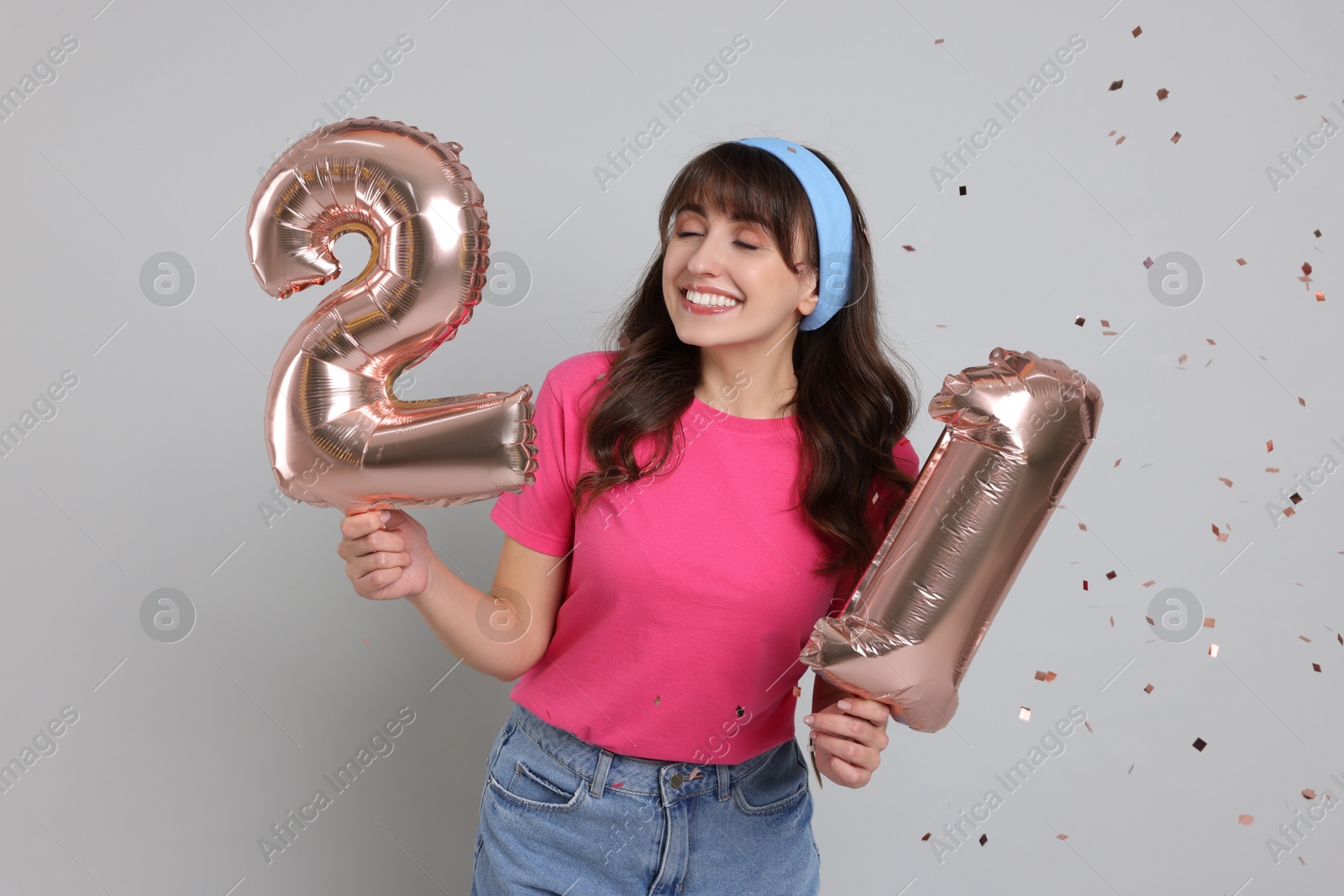 Photo of Coming of age party - 21st birthday. Young woman holding number shaped balloons on light grey background with confetti