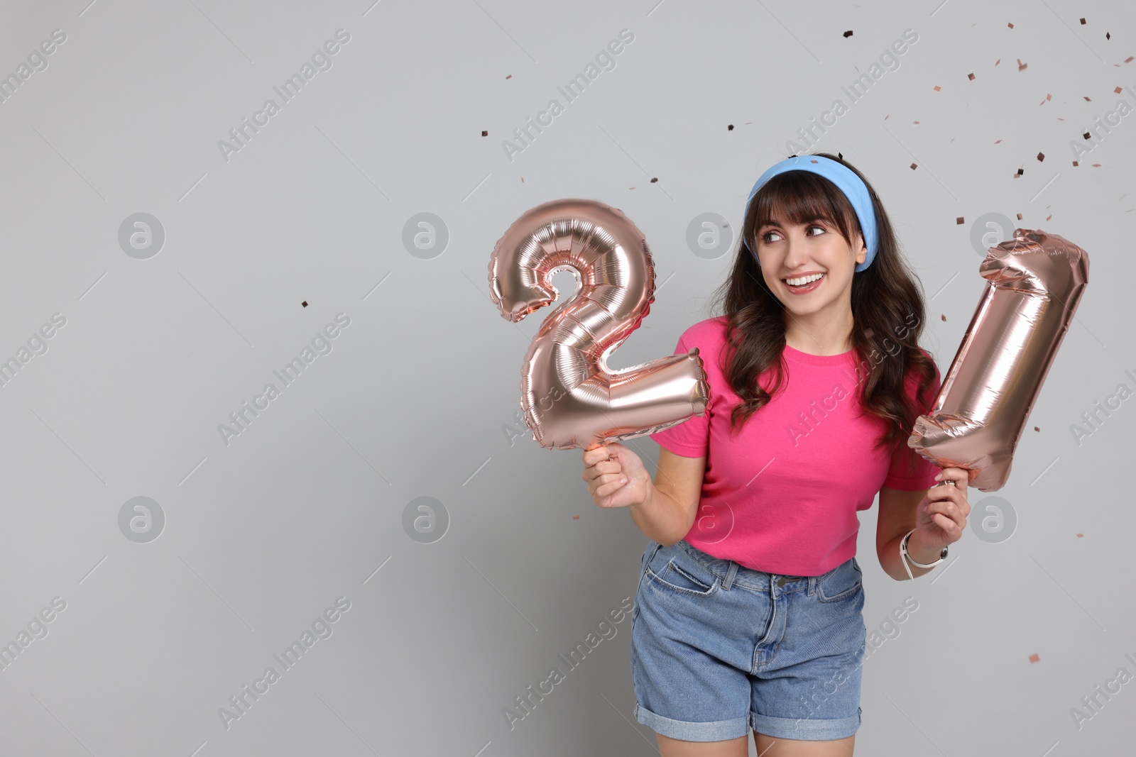 Photo of Coming of age party - 21st birthday. Young woman holding number shaped balloons on light grey background with confetti, space for text