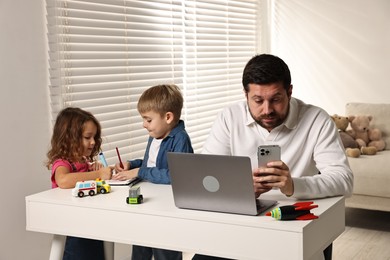 Naughty children and their overwhelmed father at table with laptop indoors