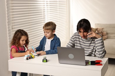 Photo of Naughty children drawing while their overwhelmed mother talking on smartphone at table indoors