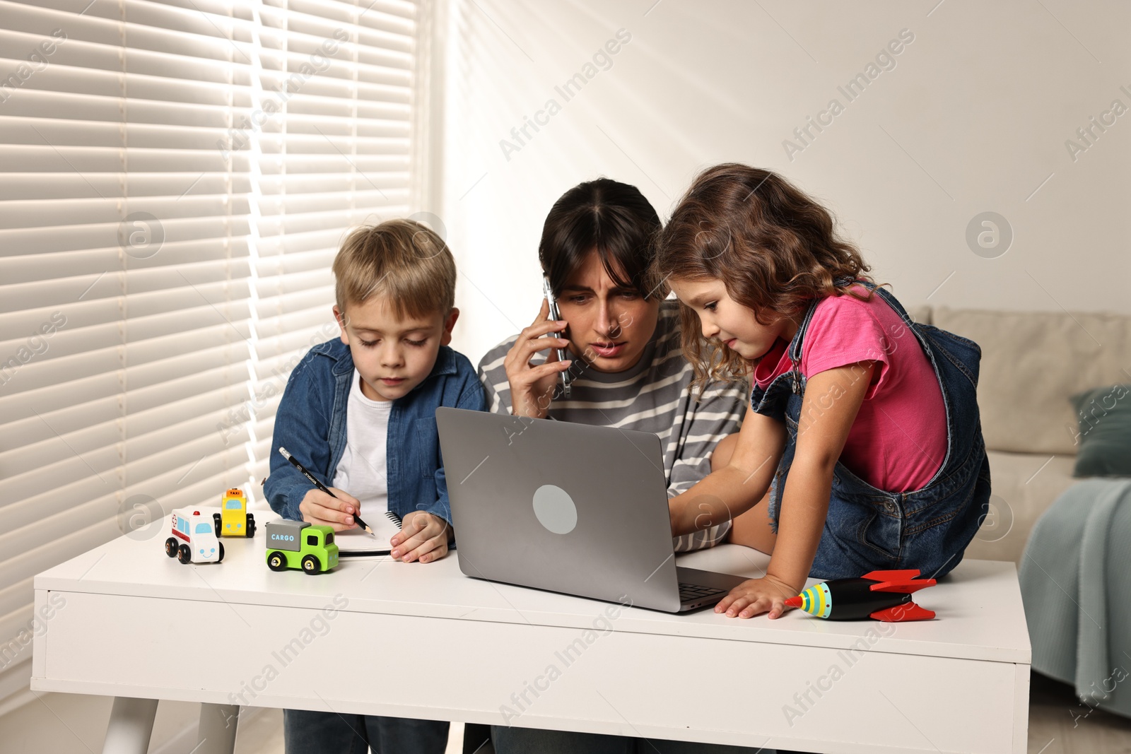 Photo of Naughty children disturbing overwhelmed mother while she talking on smartphone at table indoors