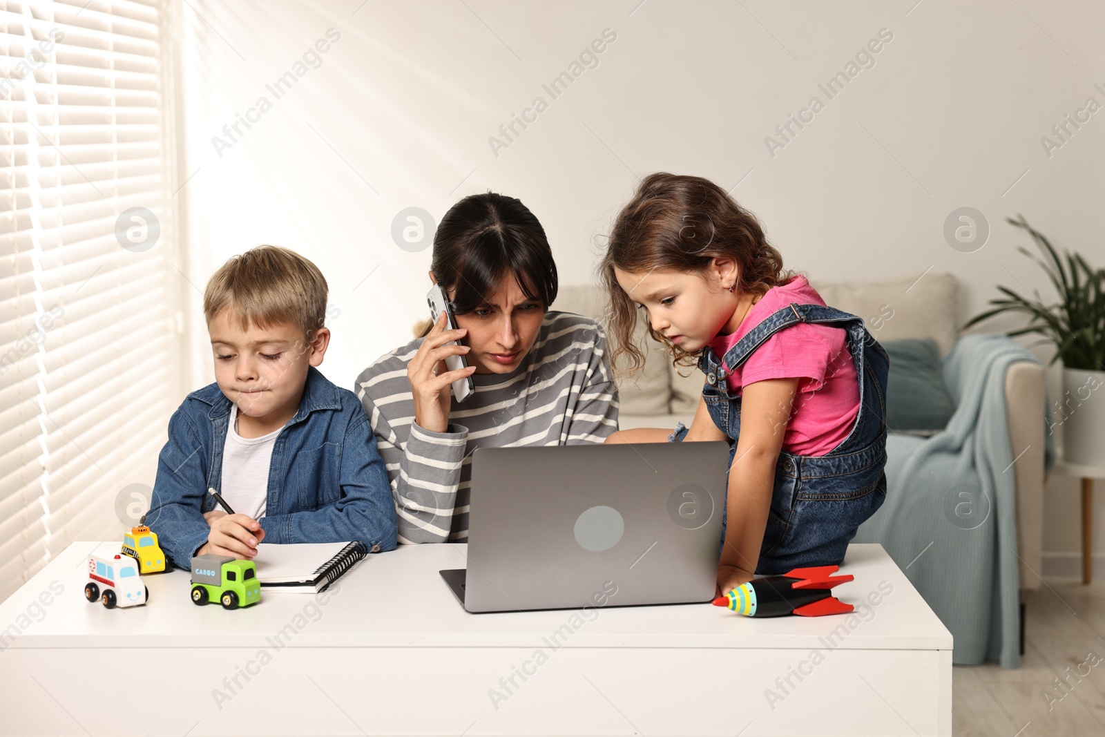 Photo of Naughty children disturbing overwhelmed mother while she talking on smartphone at table indoors