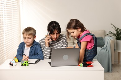 Naughty children disturbing overwhelmed mother while she talking on smartphone at table indoors