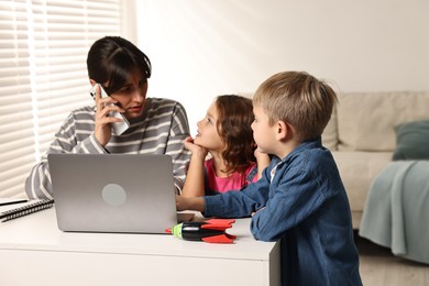 Photo of Naughty children disturbing overwhelmed mother while she talking on smartphone at table indoors