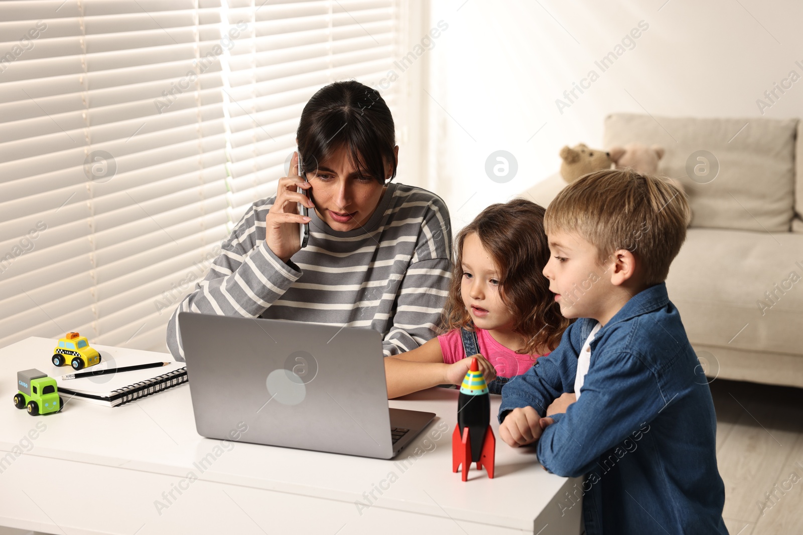 Photo of Naughty children disturbing overwhelmed mother while she talking on smartphone at table indoors