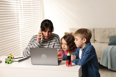 Photo of Naughty children disturbing overwhelmed mother while she talking on smartphone at table indoors