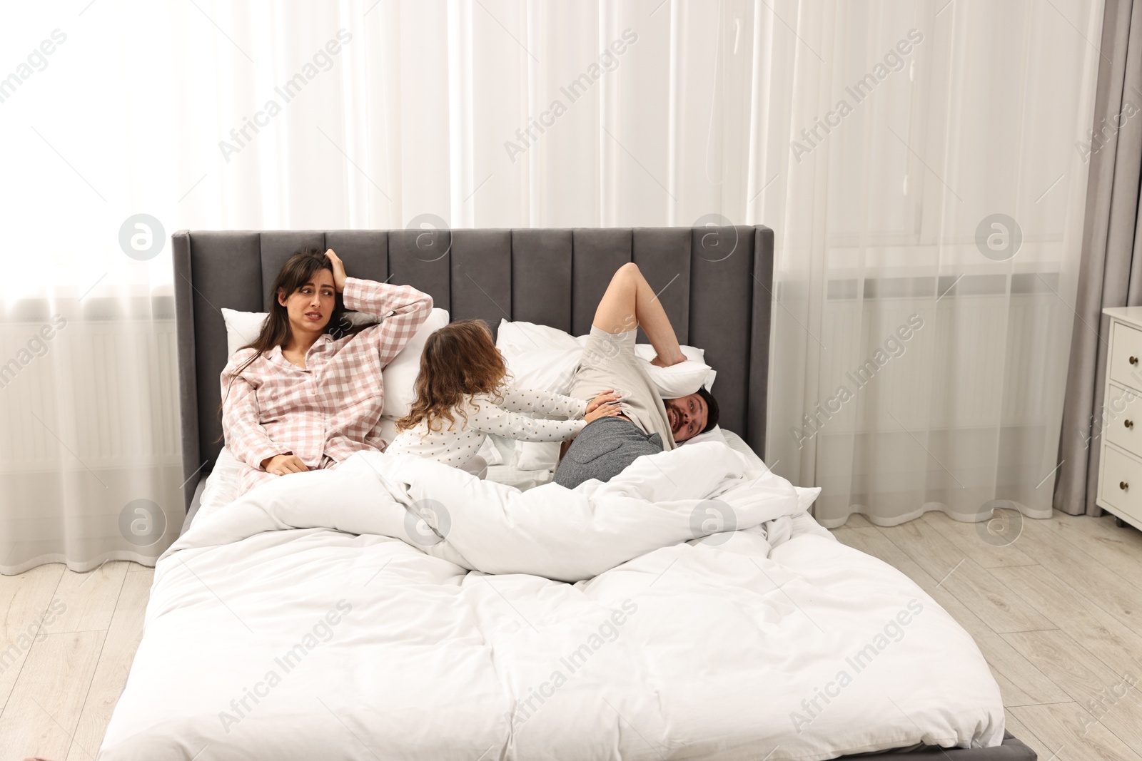 Photo of Playful daughter with her overwhelmed parents in bed at home