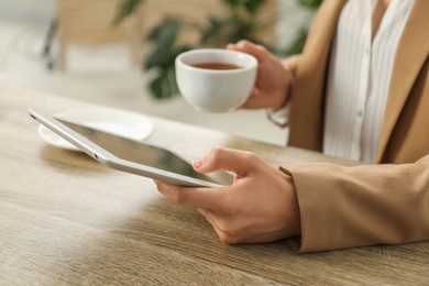 Photo of Businesswoman with cup of tea using tablet at wooden table indoors, closeup. Modern technology