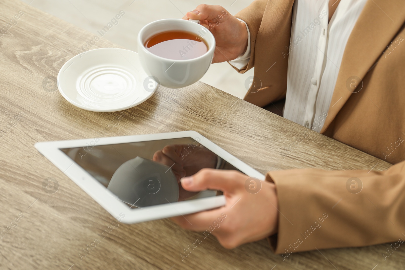 Photo of Businesswoman with cup of tea using tablet at wooden table indoors, closeup. Modern technology