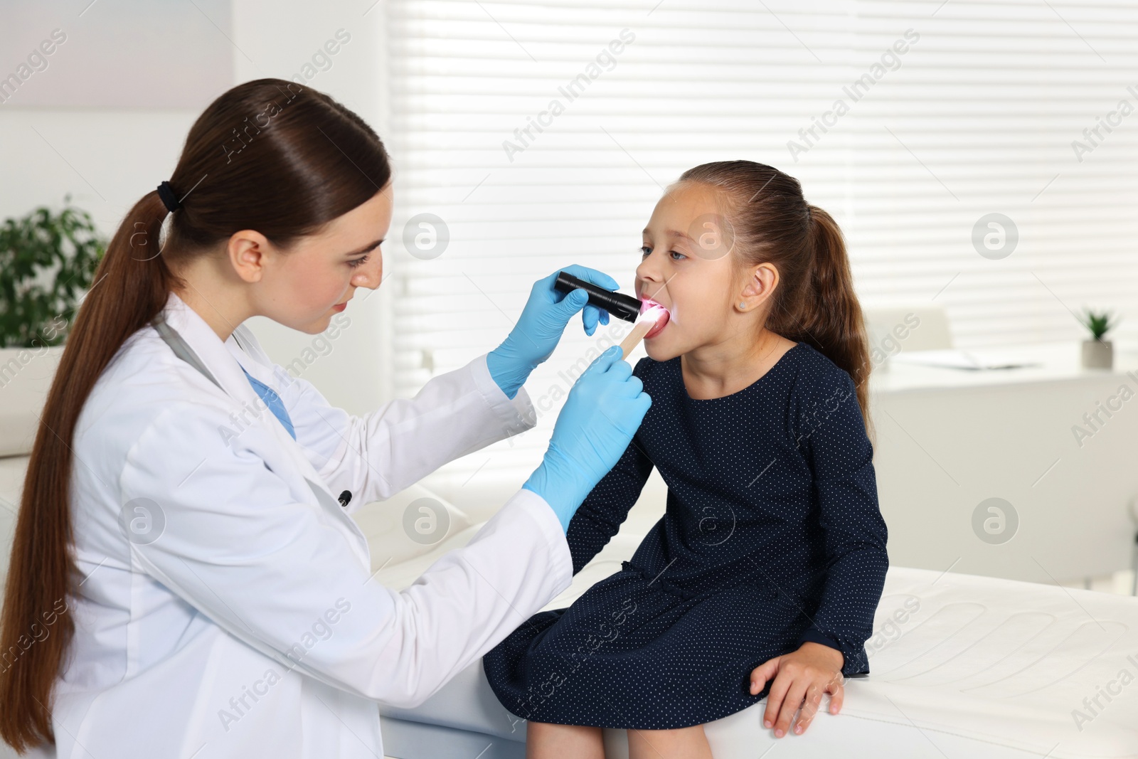 Photo of Doctor examining girl's throat with flashlight and tongue depressor in clinic